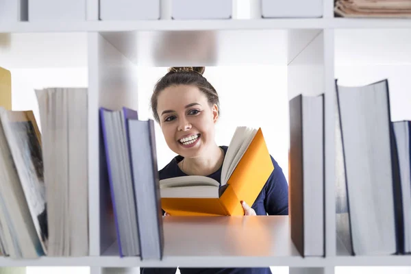 Portret van een vrolijke tiener meisje in een bibliotheek. Ze is glimlachend en een oranje boek lezen. Concept van onderwijs. — Stockfoto