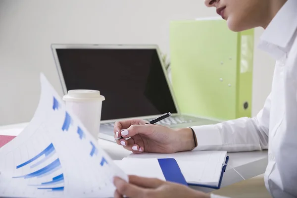 Rückansicht einer nachdenklichen, braunhaarigen Geschäftsfrau in weißer Bluse, die an ihrem Tisch in einem Büro sitzt, einen Stift in der Hand hält und ihre Notizen in einem Klemmbrett betrachtet. — Stockfoto