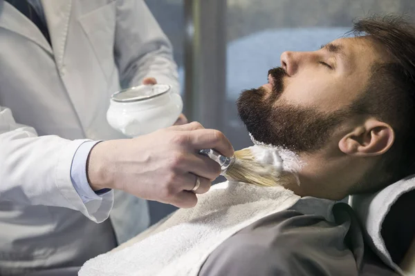 Close up of a bearded brown haired businessman sitting at a barber shop in a chair with a soap being applied to his face. — Stock Photo, Image
