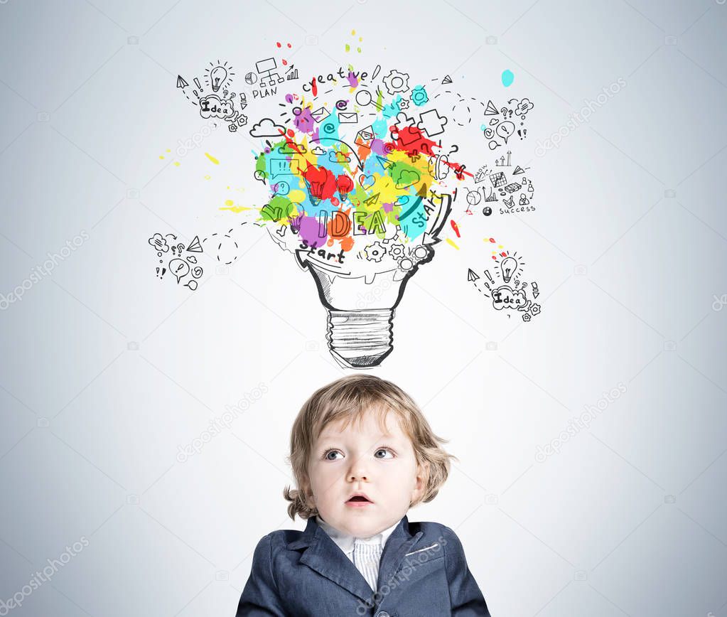 Portrait of an adorable baby boy wearing a suit and standing near a gray wall with a large and colorful start up sketch and a light bulb.