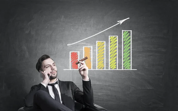 Confident european man with phone and cigar on chalkboard background with colorful business chart. Finance concept Stock Picture