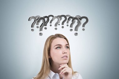 Dalgın sarışın kız ve soru işaretleri gri duvar.