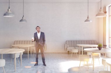 beton duvarlar, ahşap zemin, yuvarlak masa ve sandalyeler ve uzun boylu windows yakınındaki bej kanepe ile modern bir kafede Afro-Amerikan bir adamın önünde görünümü.