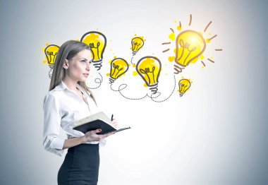 Beyaz bir bluz, siyah etek ve yüksek topuklu ayakkabı giyen ve bir defterini tutan bir sarışın iş kadını yan görünüm. Sarı ışık Ampüller beton duvara. Bir fikir kavramı