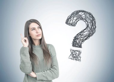 Düşünceli genç kadın uzun siyah saçlı bir yeşil kazak giyen ve yukarı doğru parmağıyla işaret. Büyük bir soru işareti ile bir gri duvar arka plan