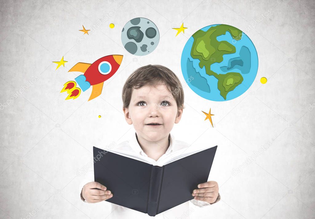 Cute little boy in a white shirt holding a book standing near a concrete wall with bright rocket, Earth and Moon drawings on it.Concept of reading and imaginary worlds.