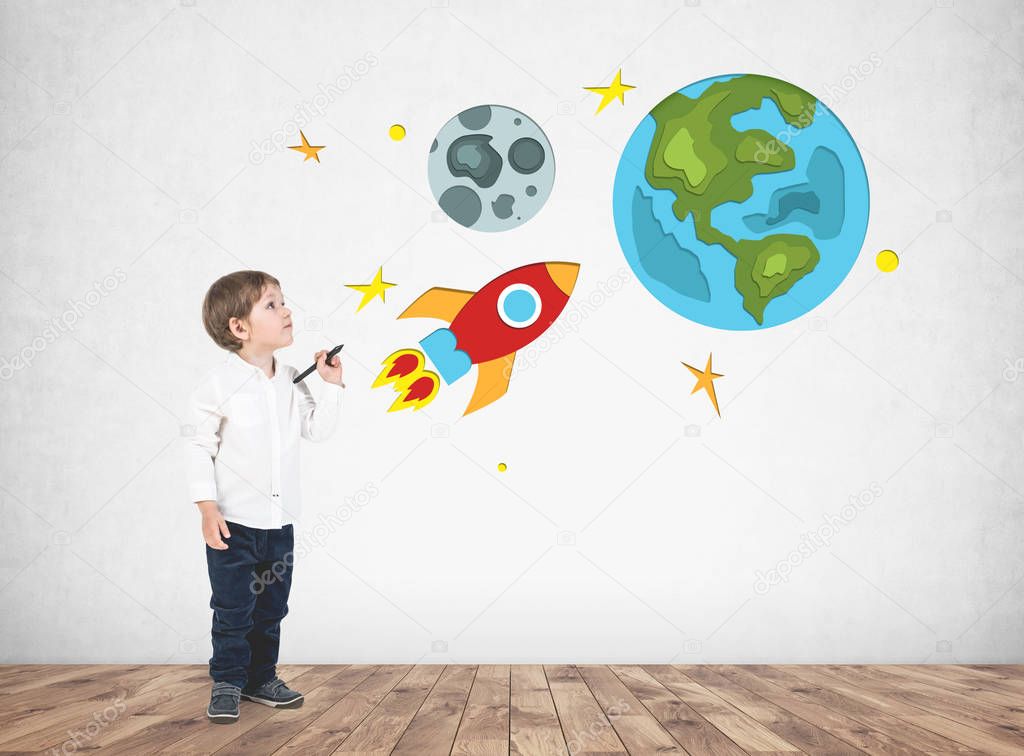 Cute little boy in a white shirt holding a marker standing near a concrete wall with bright rocket, Earth and Moon drawings on it. Concept of dreaming and imaginary worlds.