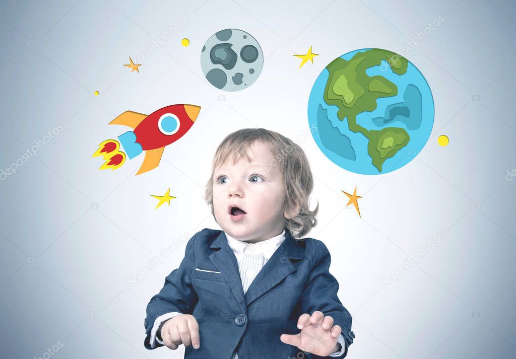 Cute astonished little boy in a suit standing near a gray wall with bright rocket, Earth and Moon drawings on it. Concept of dreaming and imaginary worlds.