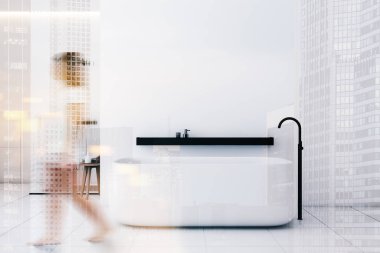 Kadın küvetli ve duşlu beyaz banyoda yürüyor.