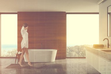 Geniş banyoda, beyaz ve ahşap duvarları, beton zemini, rahat bir küveti ve çift lavabosu olan gecelikli genç bir kadın. Deniz manzaralı pencereler. Renkli resim çift pozlama