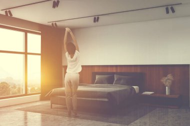 Geniş güneşli yatak odasında beyaz ve ahşap duvarlar, beton zemin, rahat bir yatak ve büyük bir pencere içinde duran pijamalı genç bir kadın. Renkli resim çift pozlama