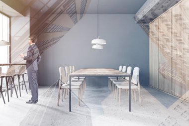 Gri ve ahşap duvarları, taş zemini, sandalyeli uzun yemek masası ve pencere kenarındaki tabureli masası olan modern restorantta kahve içen genç işadamı. Renkli resim çift pozlama