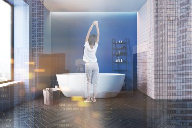 Modern banyoda mavi ve bej duvarları, siyah ahşap zemini ve rahat küveti olan pijamalı genç bir kadın. Spa konsepti. Renkli resim çift pozlama