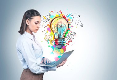 Üzerinde renkli iş fikri çizimi olan gri duvarın yanında dizüstü bilgisayarı olan ciddi genç bir kadının yan görüntüsü. Başlangıç ve yaratıcılık kavramı. Model