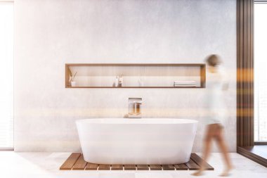 Geniş banyosunda beyaz duvarları, fayanslı zemini ve üzerinde raf olan rahat beyaz küveti olan bulanık genç bir kadın. Spa konsepti. Tonu ayarlanmış resim