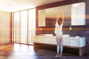 Gri duvarları, koyu fayanslı zemini, çift yuvarlak lavabosu ve yatay aynası olan lüks banyoda duran pijamalı genç bir kadın. Tonu ayarlanmış resim