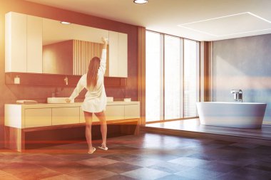 Geniş bir banyoda, gri ve ahşap duvarları, koyu fayanslı zemini, rahat bir küveti ve çift katlı lavabosu olan genç bir kadın. Tonu ayarlanmış resim