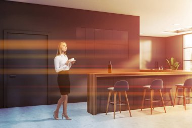 Gri duvarları, beton zemini, bar tezgahı ve mavi tabureleri olan modern bir barda kahve içen düşünceli sarışın bir iş kadını. Tonu ayarlanmış resim