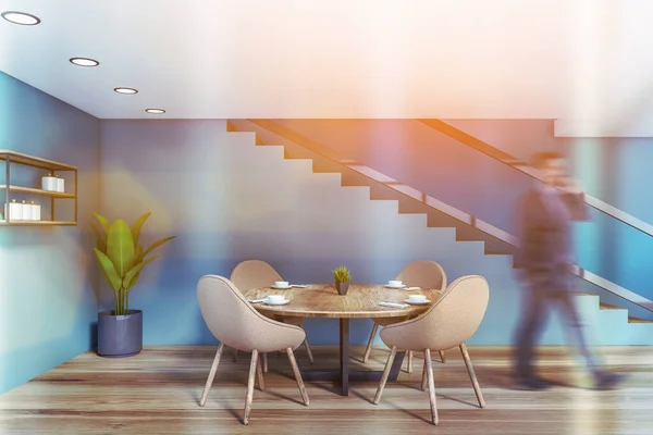 Blurry businessman on phone walking in stylish dining room with blue walls, wooden floor, round table with beige chairs and staircase. Toned image