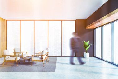 Panoramik ofis bekleme odasında gri ve ahşap duvarlar, beton zemin ve kahve masasının yanındaki beyaz koltuklar ile yürüyen bulanık genç işadamı. Tonu ayarlanmış resim