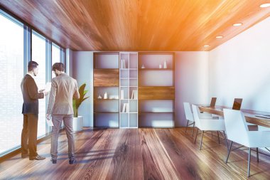 İki genç iş adamı birlikte çalışma tarzıyla birlikte beyaz açık uzay ofisinde çalışmayı tartışıyor. Uzun bilgisayar masası ve kitaplık. Renkli resim, bulanık şehir manzarası