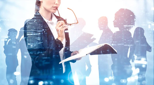 Unrecognizable young businesswoman with notebook and glasses standing over blurry night cityscape background with double exposure of her diverse business team. Concept of leadership. Toned image