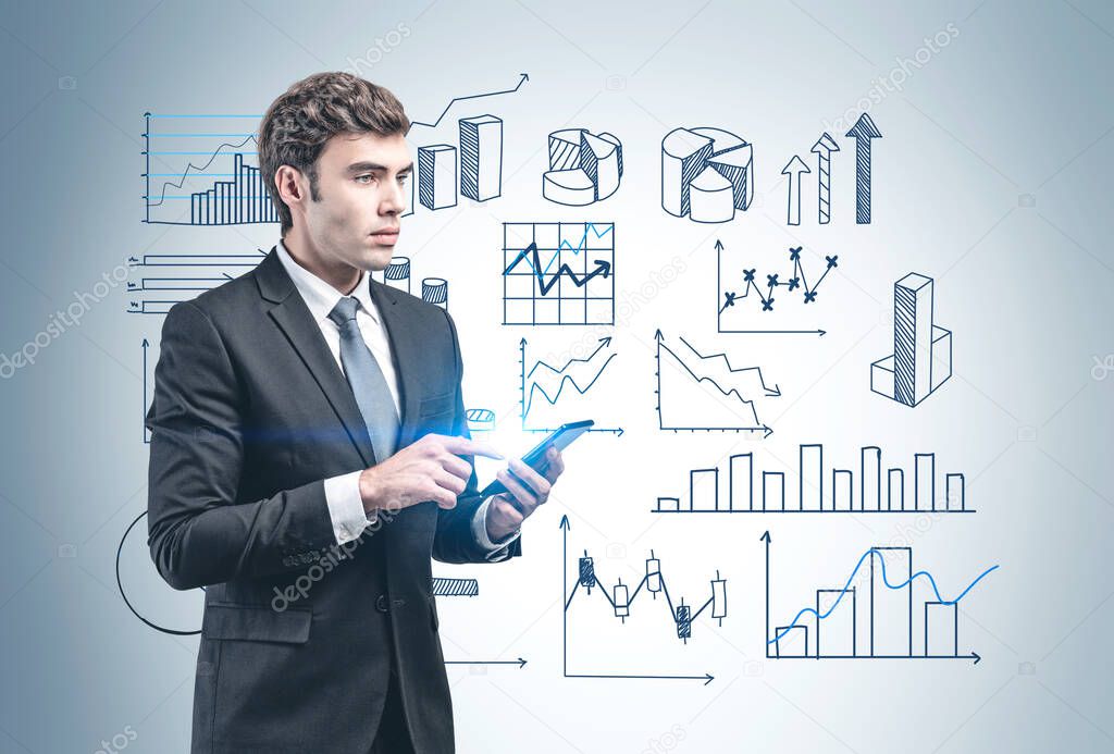 Handsome young businessman with smartphone standing near grey wall with graphs drawn on it. Concept of statistics, business education and stock market analysis