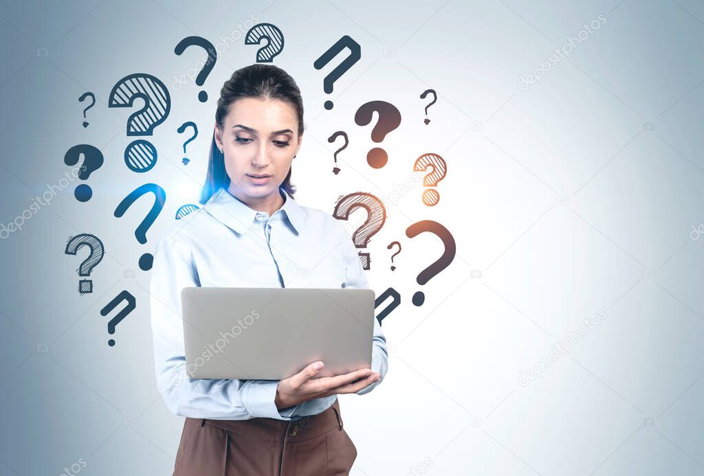 Thoughtful young businesswoman using laptop standing near grey wall with question marks drawn on it. Concept of internet search and curiosity. Mock up