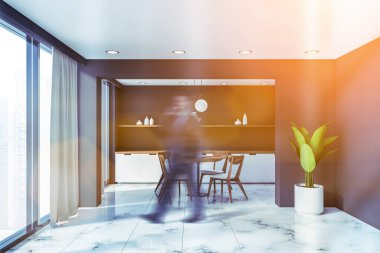 Gri duvarları, beyaz mermer zemini, beyaz tezgahları ve sandalyeli yuvarlak yemek masası olan lüks panoramik mutfakta yürüyen bulanık genç adam. Tonu ayarlanmış resim