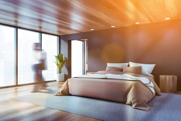グレーの壁 木製の床とベージュの毛布と快適なキングサイズベッド付きのモダンなパノラマのベッドルームで歩く若い男をブリュリー トーン画像 — ストック写真