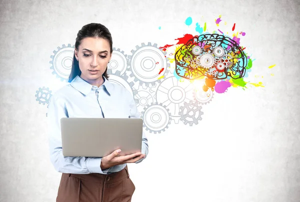 有思想的年轻女商人用笔记本电脑站在混凝土墙边 上面画着五颜六色的人脑草图 上面画着齿轮 头脑风暴和创造力的概念 — 图库照片