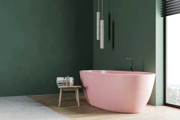 Corner of modern loft bathroom with green walls, wooden floor, comfortable pink bathtub and wooden chair with towels and beauty products. Window with blurry nice view. 3d rendering