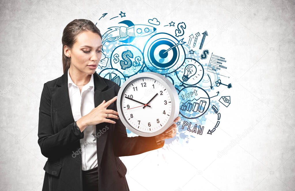 Beautiful young businesswoman holding big clock standing near concrete wall with creative startup sketch drawn on it. Concept of new project launch and time management