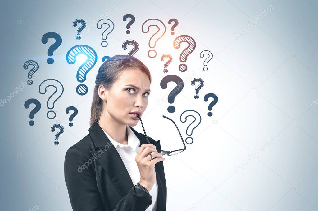 Pensive young businesswoman or college student with glasses standing near grey wall with question marks drawn on it. Concept of education and choice. Mock up