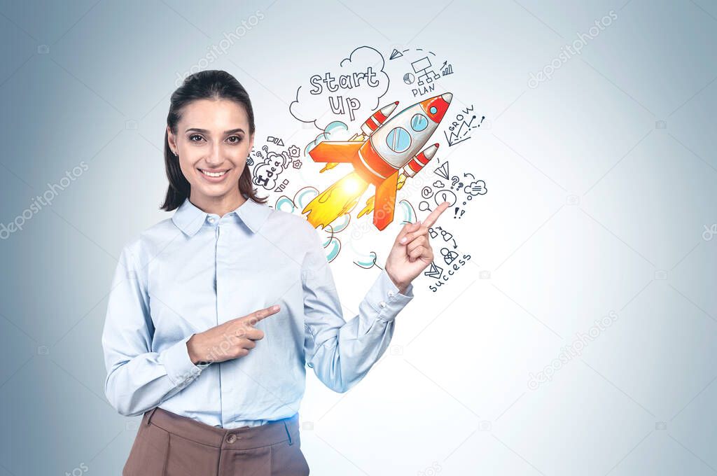 Cheerful young businesswoman with dark hair pointing at colorful startup sketch drawn on grey wall. Concept of new project launch. Mock up