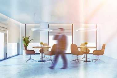 Modern ofis salonunda ya da panoramik pencereleri, beyaz duvarları ve bej koltuklu yuvarlak masaları olan bir kafede yürüyen bulanık genç işadamı. Tonu ayarlanmış resim