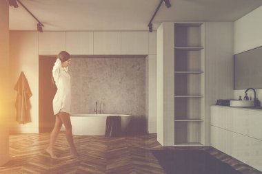 Beyaz ve beton duvarları, ahşap zemini, rahat küveti ve beyaz rafların yanında yuvarlak lavabosu olan güneş ışığında yürüyen genç bir kadın. Renkli resim çift pozlama