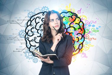 Üzerinde yaratıcı ve analitik beyin çizimi olan gri duvarın yanında duran kalem ve not defteriyle düşünceli genç iş kadını..