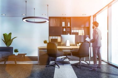 Modern CEO ofisinde beyaz duvarları, panoramik penceresi, bilgisayar masası, kitaplığı ve kanepesi olan kendinden emin genç bir iş adamı. Liderlik kavramı. Tonu ayarlanmış resim