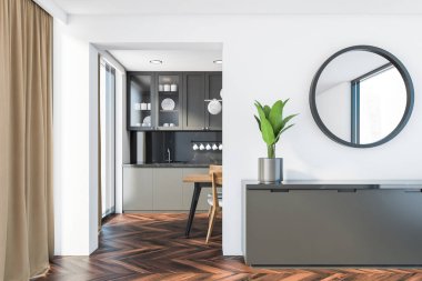 Beyaz duvarlı modern oturma odasının içi, koyu ahşap zemin, gri dolabın üstünde asılı yuvarlak ayna ve arka planda yemek masası olan lüks mutfak. 3d oluşturma