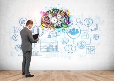 Üzerinde renkli beyin çizimi ve iş simgeleri olan beton duvarın yanında pano okuma raporu olan bir iş adamı. Beyin fırtınası kavramı