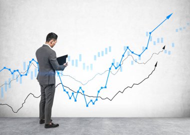 Serious young businessman reading financial report standing near concrete wall with growing graphs drawn on it. Concept of business growth and success clipart