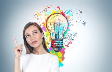 Üzerinde renkli iş fikri çizimi olan gri duvarın yanında duran uzun sarı saçlı Avrupalı genç bir kadın. Yaratıcılık ve beyin fırtınası kavramı