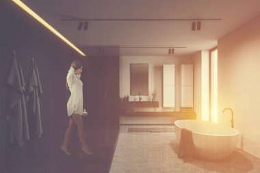 Beyaz ve gri duvarları, beton zemini, rahat küveti, ayna ve duş kabini olan güneşli banyoda yürüyen genç bir kadın. Renkli resim çift pozlama