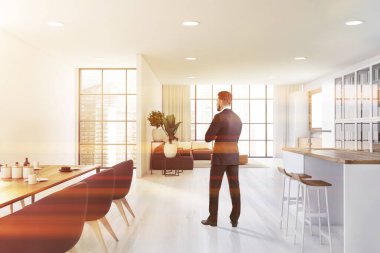 Takım elbiseli genç adam panoramik açık uzay mutfağında beyaz duvarlı, tabureli bar, koltuklu yemek masası ve arka planda gri kanepeli oturma odasında duruyor. Bulanık şehir. Tonu ayarlanmış resim