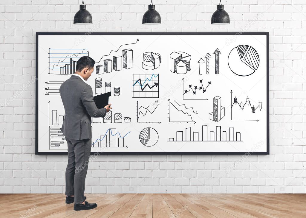 Serious young businessman reading financial report standing near whiteboard with types of graphs drawn on it. Concept of business education and statistics