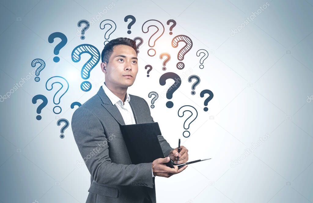 Portrait of inspired young Asian businessman writing on clipboard standing near grey wall with question marks written on it. Concept of problem solving and creativity. Mock up