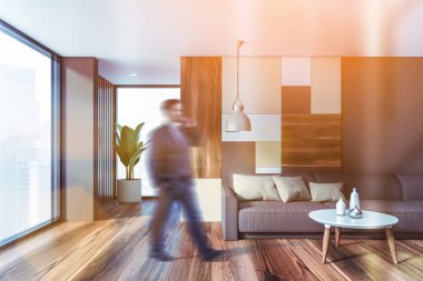 Gri ve parlak geometrik desenli duvarları, ahşap zemini ve sehpanın yanındaki rahat gri koltuğu olan şık oturma odasında yürüyen takım elbiseli bulanık genç adam. Tonu ayarlanmış resim