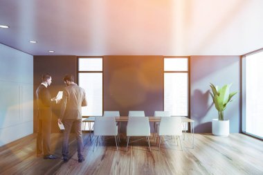 İki genç yönetici, modern toplantı odasında, gri ve beyaz duvarları olan, ahşap zemin ve beyaz koltuklu uzun konferans masası olan belgeleri tartışıyor. Tonu ayarlanmış resim