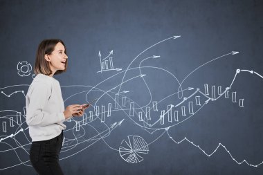 Akıllı telefonlu mutlu Avrupalı genç kızın portresi tahtaya yakın duruyor ve üzerinde bulanık mali grafikler çizilmiş. İş eğitimi ve borsa kavramı.